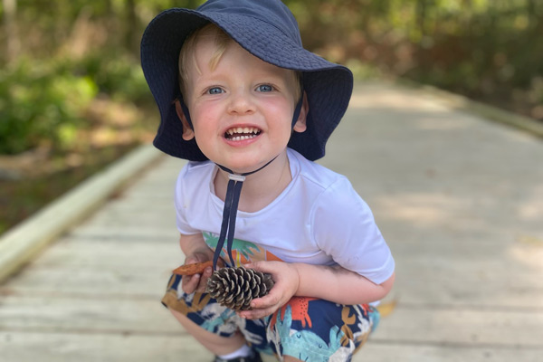Young boy in hat smiling and holding a pine cone